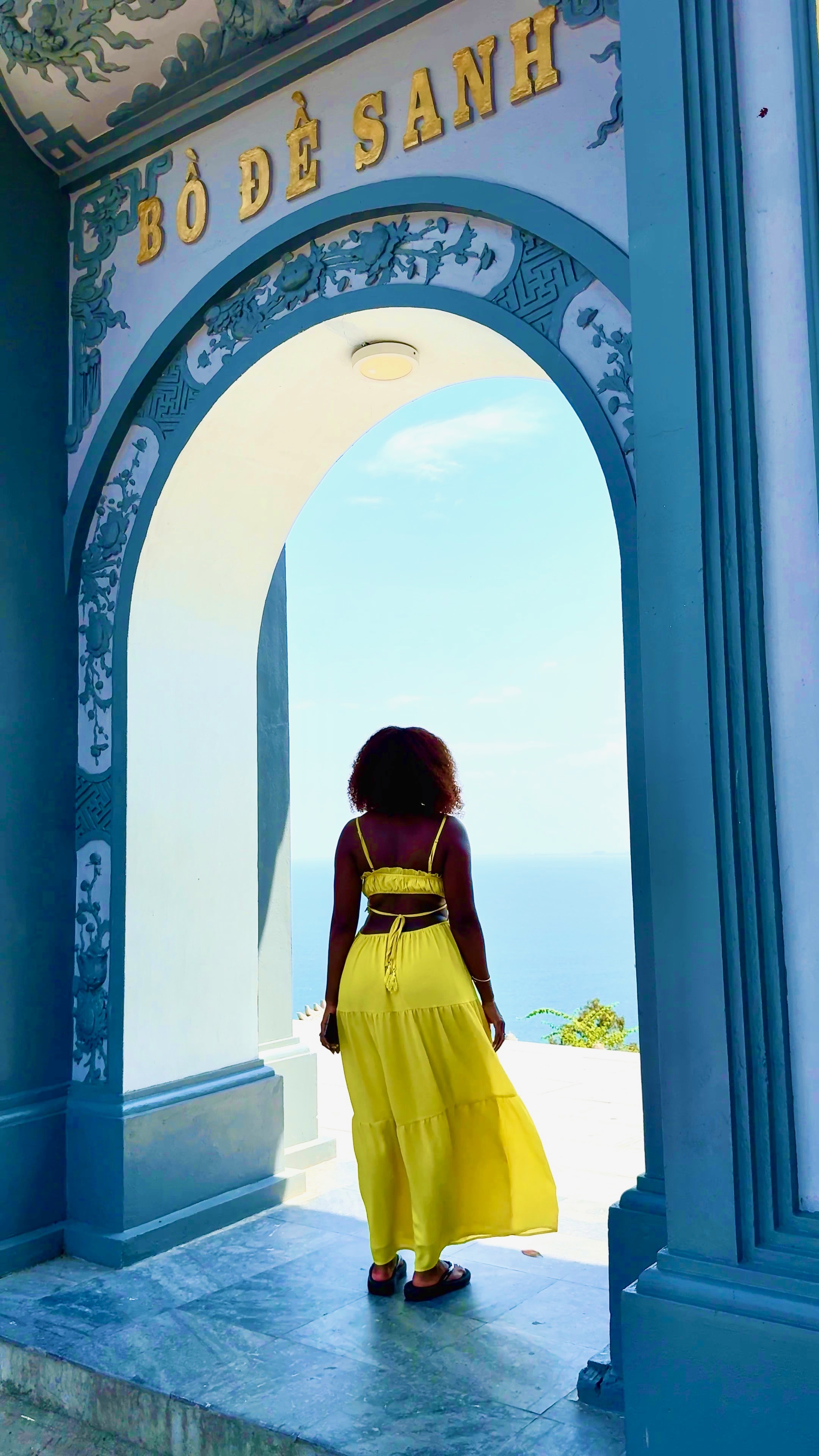Renata in a yellow dress beneath a blue archway overlooking the sea